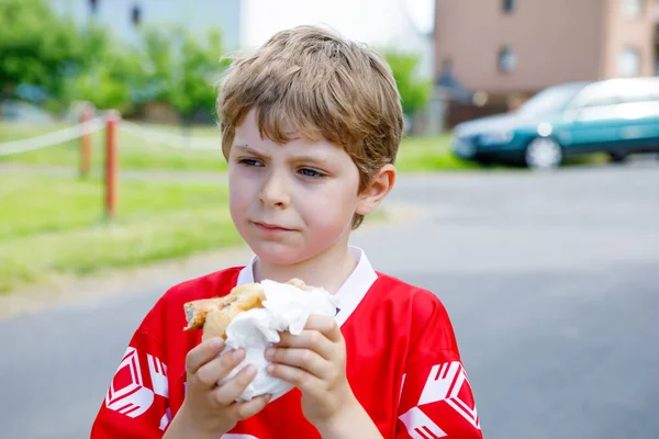 Kleine blonde jongen jongen hotdog eten na het spelen van voetbal. — Stockfoto