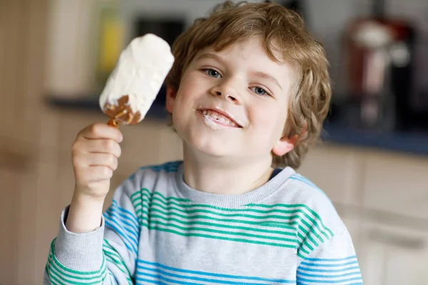 Little blond kid boy with curly hairs eating ice cream popsicle with chocolate at home — Stock Photo, Image