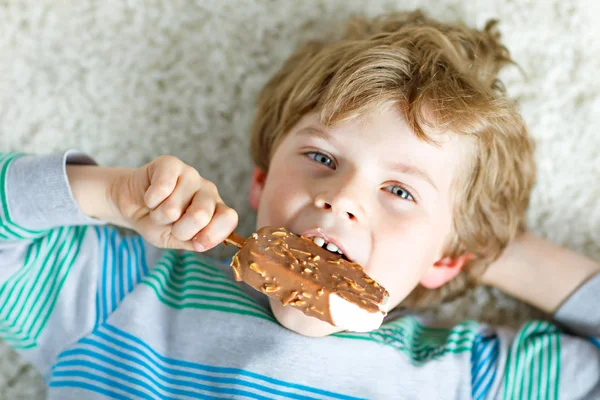 Menino loiro com cabelos encaracolados comendo gelado picolé com chocolate em casa — Fotografia de Stock