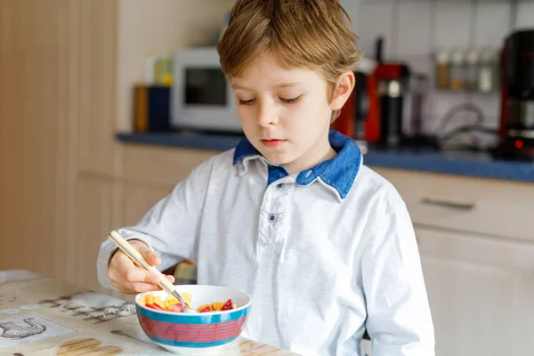 Liten blond skola unge pojke äter flingor med mjölk och bär, färska jordgubbar till frukost — Stockfoto