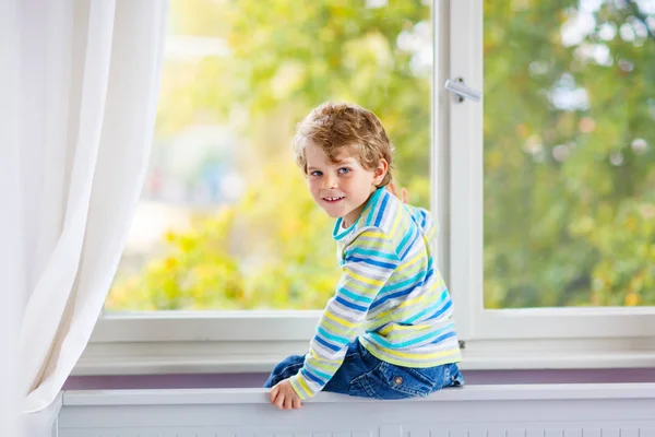 Pequeño niño sentado cerca de la ventana y mirando a los árboles de otoño —  Fotos de Stock