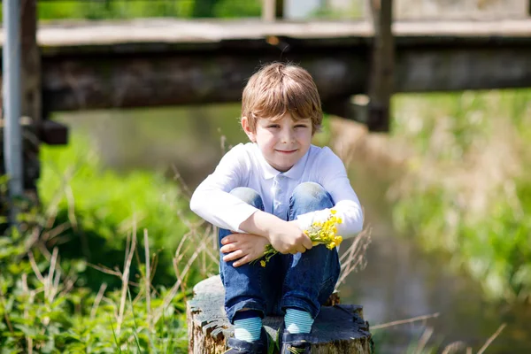 Μικρό παιδί σχολείο αγόρι κάθεται κοντά στο ποτάμι με λουλούδια στα χέρια. — Φωτογραφία Αρχείου
