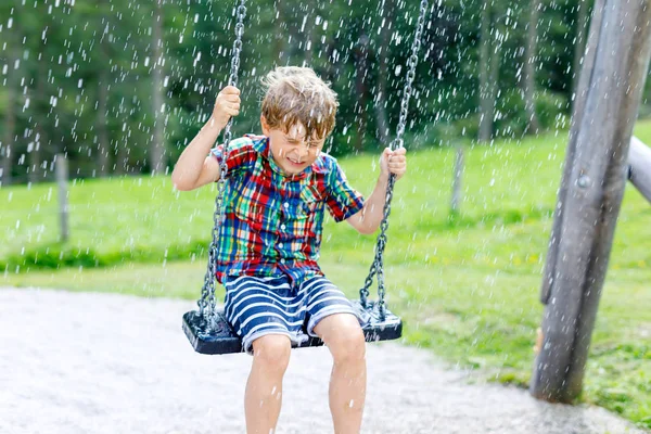 Funny kid boy having fun with chain swing on outdoor playground during rain — Stock Photo, Image