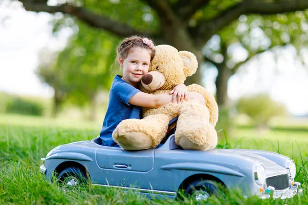 Pequena menina criança pré-escolar dirigindo carro de brinquedo grande e se divertindo com brincar com grande urso de brinquedo de pelúcia — Fotografia de Stock