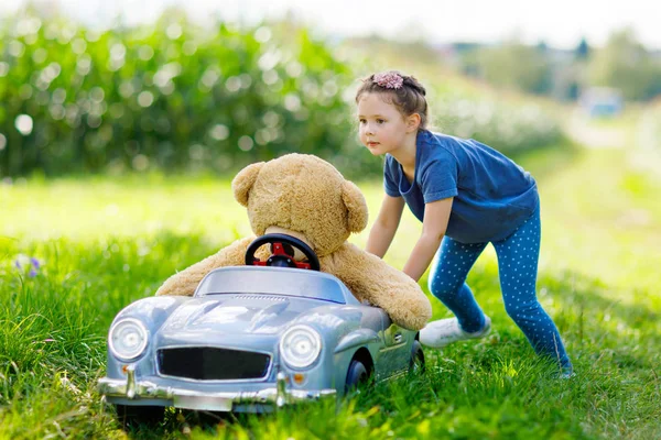就学前の小さな子供女の子大きなおもちゃの車を運転し、大きなぬいぐるみの熊と遊んで楽しい時を過す — ストック写真