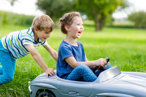 Dos niños felices niño y niña jugando con un gran coche de juguete viejo en el jardín de verano, al aire libre — Foto de Stock