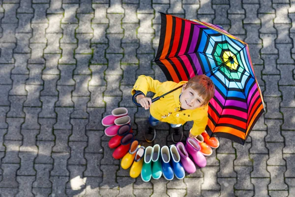 Kleiner Junge und eine Gruppe bunter Regenstiefel. Blondes Kind steht unter Regenschirm. — Stockfoto