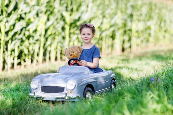 Pequena menina criança pré-escolar dirigindo carro de brinquedo grande e se divertindo com brincar com grande urso de brinquedo de pelúcia — Fotografia de Stock