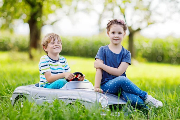 Двое счастливых детей играют с большой старой игрушечной машиной в летнем саду, на открытом воздухе — стоковое фото