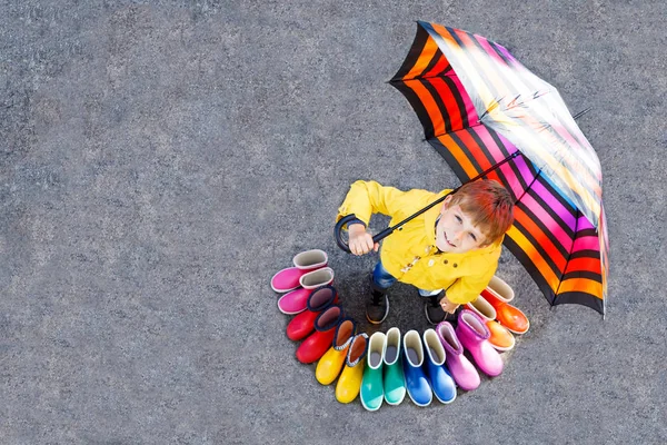 Ragazzino e gruppo di stivali da pioggia colorati. Biondo bambino in piedi sotto l'ombrello . — Foto Stock