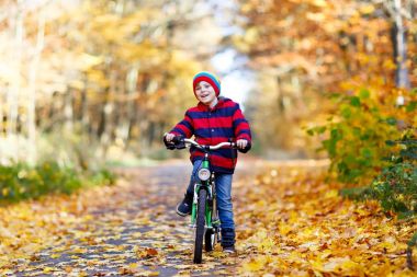 Sonbahar orman parkında bisiklet süren, renkli, sıcacık elbiseli bir çocuk.