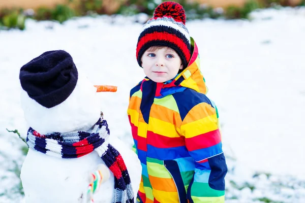 Funny kid boy in colorful clothes making a snowman, outdoors — Stock Photo, Image