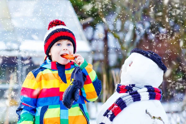 Смешной мальчик в разноцветной одежде делает снеговика на открытом воздухе — стоковое фото