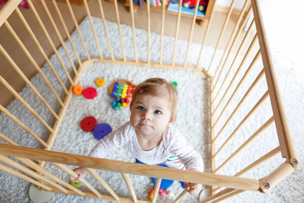 Beautiful little baby girl standing inside playpen. Cute adorable child playing with colorful toy — Stock Photo, Image