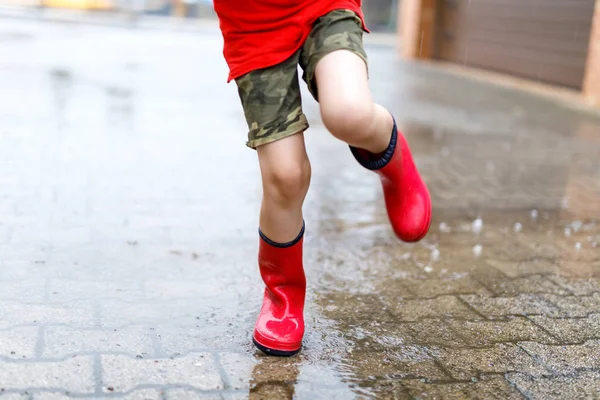 Criança usando botas de chuva vermelha pulando em uma poça . — Fotografia de Stock