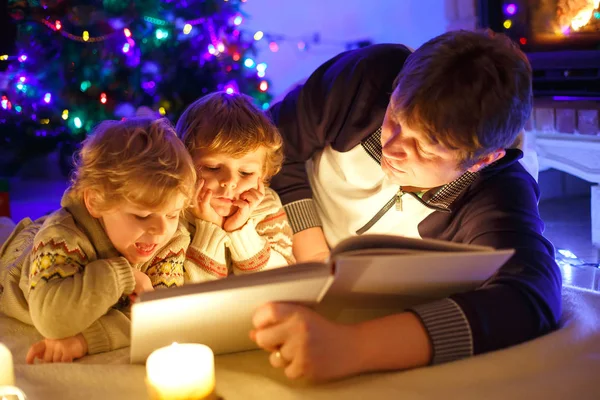 Vater und zwei kleine Jungen lesen Buch am Kamin, Kerzen und Kamin. — Stockfoto