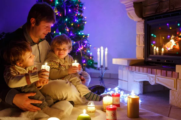 Vater und zwei kleine Jungen sitzen am Kamin, Kerzen und Kamin und schauen auf das Feuer. — Stockfoto