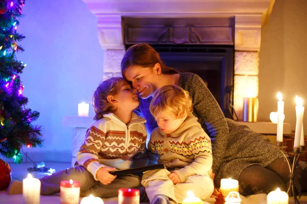 Ung mamma och hans två små barn sitter en öppen spis hemma på jul — Stockfoto