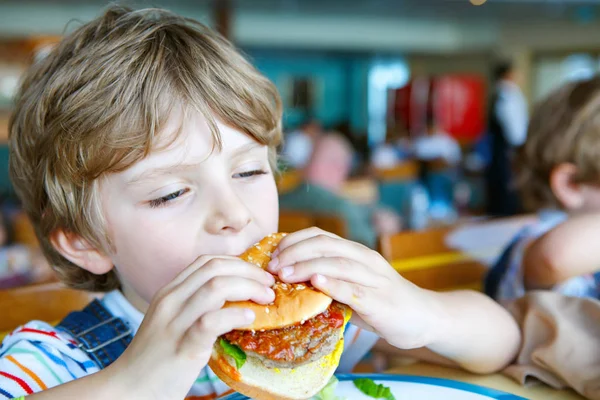 Cute healthy preschool boy eats hamburger sitting in school canteen — Stock Photo, Image