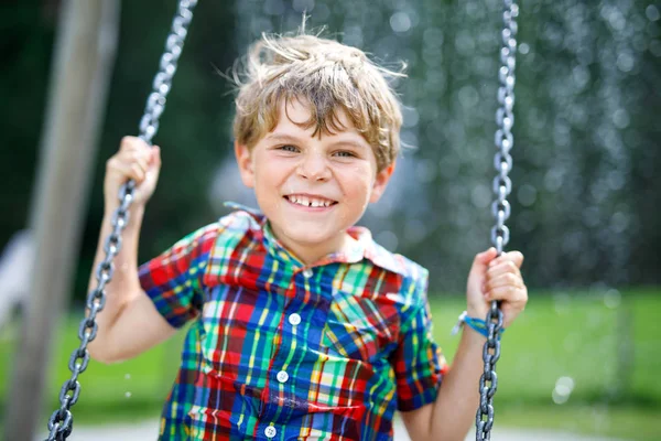 Lustiger Junge hat bei Regen Spaß mit Kettenschaukel auf Spielplatz im Freien — Stockfoto