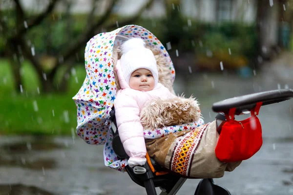 Schattig klein mooie babymeisje, zittend in de kinderwagen of wandelwagen op koude dag met regen, ijzel en sneeuw. — Stockfoto