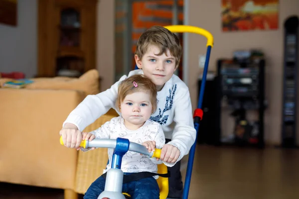 Niño pequeño empujando bicicleta o triciclo con hermana pequeña linda. Niña y hermano jugando juntos en casa, adentro . — Foto de Stock