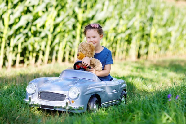 Pequena menina criança pré-escolar dirigindo carro de brinquedo grande e se divertindo com brincar com grande urso de brinquedo de pelúcia — Fotografia de Stock