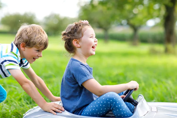 Двое счастливых детей играют с большой старой игрушечной машиной в летнем саду, на открытом воздухе — стоковое фото