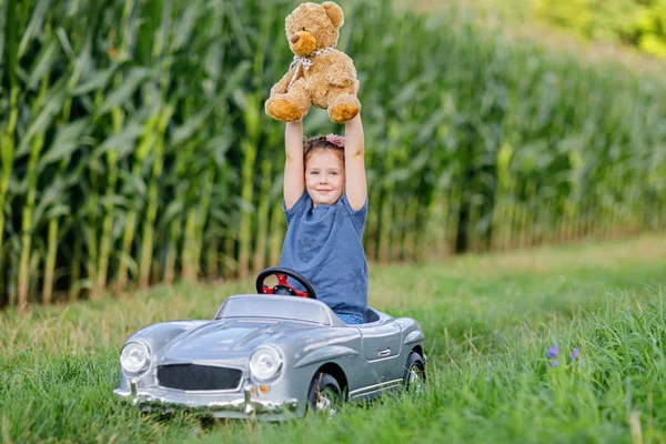 Pequeña niña preescolar conduciendo un gran coche de juguete y divirtiéndose jugando con un gran oso de peluche — Foto de Stock