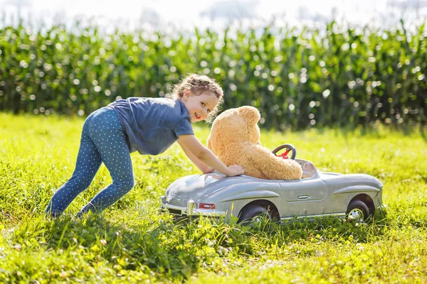 Pequena menina criança pré-escolar dirigindo carro de brinquedo grande e se divertindo com brincar com grande urso de brinquedo de pelúcia — Fotografia de Stock