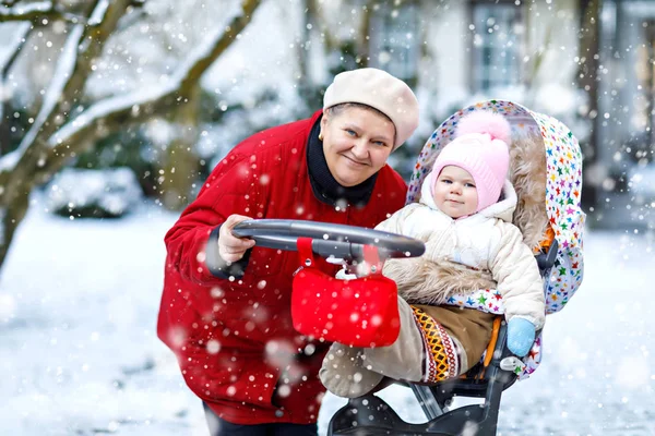 Vackra mormor promenader med baby flicka i barnvagn under snöfall på vintern — Stockfoto