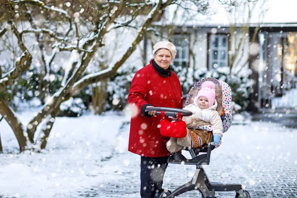 Vackra mormor promenader med baby flicka i barnvagn under snöfall på vintern — Stockfoto