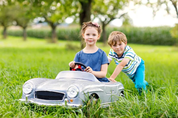 Duas crianças felizes brincando com o carro de brinquedo velho grande no jardim de verão, ao ar livre — Fotografia de Stock