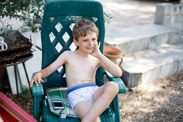 Крутой мальчик, сидящий в кресле в открытом саду с мобильным телефоном. Мальчик наслаждается отпуском — стоковое фото