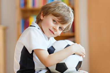 Futbol topu olan sarışın anaokulu çocuğu televizyonda futbol maçı izliyor..