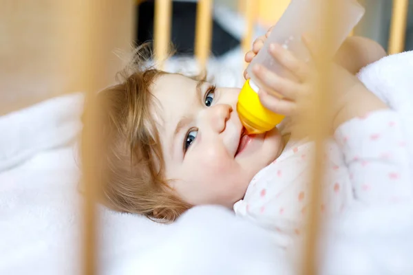 Χαριτωμένο κοριτσάκι κρατώντας μπουκάλι με φόρμουλα ήπια και πόσιμο. Παιδί σε βρεφικό κρεβάτι πριν κοιμηθεί — Φωτογραφία Αρχείου
