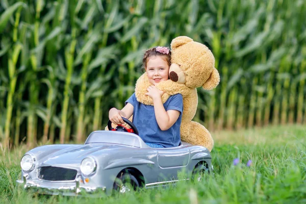 Little preschool kid girl driving big toy car and having fun with playing with big plush toy bear — Stock Photo, Image