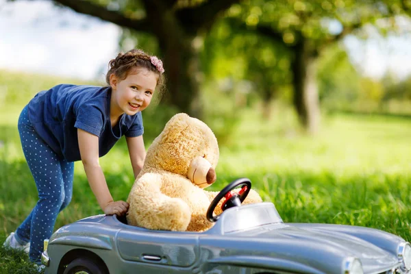 Pequeña niña preescolar conduciendo un gran coche de juguete y divirtiéndose jugando con un gran oso de peluche — Foto de Stock