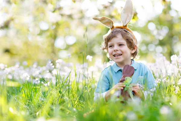 Cute little kid boy with Easter bunny ears celebrating traditional feast. Happy child eating chocolate rabbit fugure — Stock Photo, Image