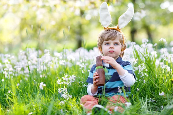 Netter kleiner Junge mit Osterhasenohren feiert traditionelles Fest. Glückliches Kind isst Schokohasen-Fugure — Stockfoto