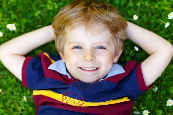 Happy little blond child, kid boy with blue eyes laying on the grass with daisies flowers in the park. — Stock Photo, Image