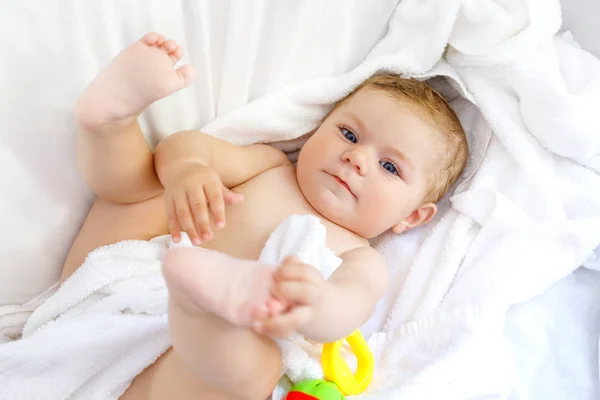 Niedliches kleines Baby, das nach dem Baden mit Spielzeugrassel und eigenen Füßen spielt. entzückende schöne Mädchen in weiße Handtücher gehüllt — Stockfoto