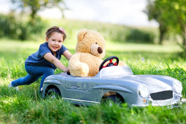 Pequeña niña preescolar conduciendo un gran coche de juguete y divirtiéndose jugando con un gran oso de peluche — Foto de Stock
