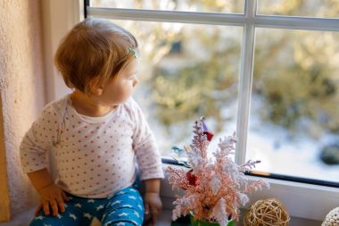 Mutlu sevimli kız bebek pencerenin yanında oturuyor ve kışın veya ilkbaharda dışarıda karları seyrediyor.