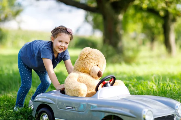 Kleines Vorschulmädchen fährt großes Spielzeugauto und hat Spaß beim Spielen mit großem Plüschbär — Stockfoto