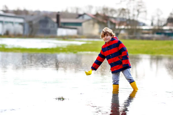 Niño feliz en botas de lluvia amarillas jugando con barco de papel por charco enorme en el día de primavera u otoño —  Fotos de Stock