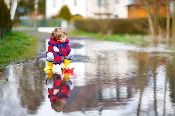 Menino pequeno feliz em botas de chuva amarela jogando com barco de navio de papel por enorme poça no dia de primavera ou outono — Fotografia de Stock