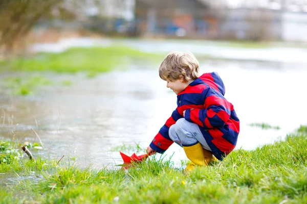 Szczęśliwy chłopiec w żółtych butach deszczowych bawiących się papierową łodzią przez ogromną kałużę wiosną lub jesienią — Zdjęcie stockowe