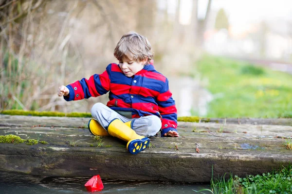 Menino pequeno feliz em botas de chuva amarela jogando com barco de navio de papel por enorme poça no dia de primavera ou outono — Fotografia de Stock