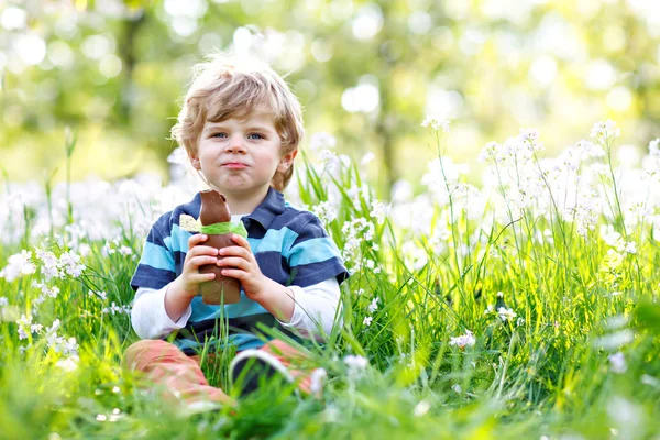 Netter kleiner Junge mit Osterhasenohren feiert traditionelles Fest. Glückliches Kind isst Schokohasen-Fugure — Stockfoto